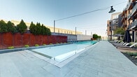 This outdoor amenity space offers a sophisticated urban retreat centered around a generously sized lap pool with crystal-clear blue water. The pool is framed by sleek tiled surrounds and equipped with entry handrails, making it both functional and visually striking. A wide, stone-paved pool deck runs alongside the water, providing ample space for relaxation and movement.

Adjacent to the pool, a warm wood-decked lounge area is lined with contemporary chaise lounge chairs and large shade umbrellas, creating an inviting setting for sunbathing and leisure. Thoughtfully placed greenery, including columnar evergreen trees and low ornamental plantings, adds a natural touch to the urban environment.

The space is enclosed on one side by a combination of weathered Corten steel panels and a textured stone accent wall, offering both privacy and an industrial-modern aesthetic. On the opposite side, a multi-story brick residential building rises alongside the deck, its large windows and private balconies overlooking the pool area. Overhead string and pendant lighting provides ambiance during evening hours. The overall design seamlessly blends modern architecture with functional outdoor living, creating a compelling amenity space suited for both active use and relaxed enjoyment.