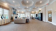 Located at 2015 Wave Crest Dr in Raleigh, NC, this single-floor clubhouse showcases refined architectural design and upscale finishes throughout. The centerpiece of the main lounge is a striking series of octagonal coffered ceilings with recessed lighting, lending the space a grand, distinguished character. The lounge area features a classic white fireplace flanked by built-in shelving, plush neutral sectional seating with teal accent pillows, and large French doors that invite abundant natural light and outdoor views.

The foyer greets visitors with soaring tray ceilings, decorative crown molding, and an elegant console table framed by tall vases and a geometric mirror wall accent. The transition into the dining area reveals wood-look tile flooring, expansive multi-pane windows with patterned valances, and a mix of dining table seating and bar-height options, making it well-suited for both casual and formal gatherings.

The kitchen is appointed with rich dark wood cabinetry, marble-look countertops, a herringbone tile backsplash, a central island with bar seating, and stainless steel appliances. Warm pendant lighting and large windows complete the functional yet stylish space. Throughout, the design balances warm neutral tones with contemporary accents, creating a cohesive and welcoming community environment.