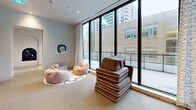 Located at 65 Broadway Ave in Toronto's midtown neighbourhood, this single-floor space offers a thoughtfully designed layout comprising a living room and a kitchenette. The living area is the standout feature of the space, defined by expansive floor-to-ceiling windows framed in black metal that flood the interior with natural light and offer open views of the surrounding urban streetscape. Elegant patterned drapes complement the clean, neutral palette of white walls and speckled light grey flooring, contributing to a calm and modern ambiance. The room accommodates a variety of flexible soft seating, reinforcing its relaxed and welcoming atmosphere. Wall-mounted artwork adds a subtle decorative touch. The kitchenette is compact and efficient, outfitted with smooth white cabinetry — both upper and lower — featuring black hardware, a stainless steel sink with a matte black faucet, and a grey countertop, all maintaining the space's clean contemporary aesthetic. Recessed lighting throughout ensures consistent illumination across both areas. Overall, the space presents a bright, well-appointed interior with a minimalist modern design suited for comfortable, everyday use.
