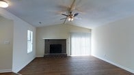 Located at 1398 S. University Drive in Ladson, South Carolina, this single-story home offers a comfortable and inviting layout with notable architectural details throughout. The living room serves as a focal point of the home, showcasing soaring vaulted ceilings that lend an open, airy feel to the space. A classic brick fireplace with a clean white mantel anchors the room, adding warmth and character. Rich, hardwood-style flooring runs throughout, complementing the neutral-toned walls and creating a cohesive, welcoming atmosphere. A ceiling fan with an integrated light fixture is centrally positioned beneath the vaulted ceiling, providing both comfort and illumination. Natural light fills the living area through a large sliding glass door dressed with vertical blinds, as well as an additional window, keeping the space bright and connected to the outdoors. The textured ceiling adds a layer of classic detail, while the overall design balances practicality with charm. This well-appointed single-floor home in Ladson offers a straightforward, functional layout ideal for comfortable everyday living.