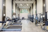 This two-story amenity facility is thoughtfully designed to support an active lifestyle, combining generous open spaces with a wide array of fitness equipment and inviting common areas. The entrance foyer greets visitors with soaring ceilings, light hardwood flooring, and a sleek staircase with black metal railings that connects both levels. Large multi-pane windows and glass doors flood the entry with natural light and offer views of the surrounding greenery.

The main fitness area is the centerpiece of the facility, boasting dramatic double-height ceilings accented by exposed steel columns and a striking arched window that frames views of the mature trees outside. It is comprehensively outfitted with a row of Matrix treadmills and ellipticals, a full selection of strength machines, free weights, cable systems, and functional training accessories including medicine balls and stability equipment. Polished concrete floors and full-length mirrors complete the professional gym aesthetic.

A secondary workout room, finished with warm hardwood flooring, provides a quieter space for rowing machines and additional cardio equipment, surrounded by floor-to-ceiling windows overlooking wooded scenery. The open lounge area features a distinctive reclaimed wood accent wall, large windows, and hardwood floors, creating a welcoming gathering space. Accessible restrooms with tile flooring are conveniently located on-site.