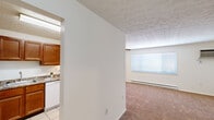 This 2-bedroom, 1-bathroom single-floor unit offers a practical and comfortable layout across a well-organized floor plan. The combined kitchen and living room serves as the heart of the home, where a carpeted living area is anchored by a ceiling fan with light fixture and a large window that allows natural light to fill the room. A baseboard heating system runs along the walls, ensuring year-round comfort. The kitchen is equipped with wood cabinetry, a white electric range, tile flooring, and a pass-through window connecting it to the living space, adding an open, communicative feel between the two areas.

The spacious primary bedroom features carpet flooring, a large window with blinds, a wall-mounted air conditioning unit, and baseboard heating. The second bedroom is similarly finished with soft carpet, a closet with louvered doors, and direct access to the hallway. Both bedrooms offer generous proportions suitable for comfortable furnishing.

The full bathroom is finished with white tile wainscoting, tile flooring, a white vanity with under-cabinet storage, and a shower-tub combo. A central hallway with dark wood doors connects the bedrooms and bathroom efficiently. Throughout the unit, textured ceilings add a classic residential character to the overall design.
