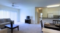 This single-floor residence offers a thoughtfully designed open-concept layout that seamlessly connects the living, dining, and kitchen areas. The living room is bright and airy, featuring plush wall-to-wall carpeting, neutral white walls, and large vertical-blind-covered windows that allow for ample natural light. A ceiling fan overhead provides comfort and airflow throughout the space. The dining area sits adjacent to the living room, accommodating a dark-wood dining set that complements the overall color palette. A breakfast bar visually separates the kitchen from the main living space while maintaining the open feel of the floor plan. The kitchen is accessible via a short corridor and features dark countertops that contrast cleanly against lighter cabinetry. Louvered bi-fold doors provide access to additional storage or utility space, adding practicality to the layout. The overall ambiance is clean, neutral, and functional, with a cohesive dark-wood furniture theme running throughout. The uncluttered design and open sightlines make the space feel expansive and easy to navigate, offering a comfortable and livable environment.