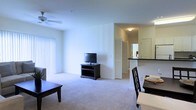 This single-floor residence offers a thoughtfully designed open-concept layout that seamlessly connects the living, dining, and kitchen areas. The living room is bright and airy, featuring plush wall-to-wall carpeting, neutral white walls, and large vertical-blind-covered windows that allow for ample natural light. A ceiling fan overhead provides comfort and airflow throughout the space. The dining area sits adjacent to the living room, accommodating a dark-wood dining set that complements the overall color palette. A breakfast bar visually separates the kitchen from the main living space while maintaining the open feel of the floor plan. The kitchen is accessible via a short corridor and features dark countertops that contrast cleanly against lighter cabinetry. Louvered bi-fold doors provide access to additional storage or utility space, adding practicality to the layout. The overall ambiance is clean, neutral, and functional, with a cohesive dark-wood furniture theme running throughout. The uncluttered design and open sightlines make the space feel expansive and easy to navigate, offering a comfortable and livable environment.