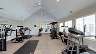 Located at 1400 Laurel Springs Dr in Durham, NC, this single-floor fitness center is designed to provide a welcoming and functional workout environment. The room is defined by its striking vaulted ceiling, which lends a sense of openness and airiness to the space. Dual ceiling fans circulate air throughout, while recessed lighting ensures the area is well-lit at all times. Large windows along the walls invite generous natural light, further enhancing the bright and energizing atmosphere. The fitness center is equipped with a comprehensive selection of exercise equipment, including treadmills, a Landice cardio machine, a multi-station cable and weight machine, a workout bench, and yoga/exercise mats for floor-based training. The light-toned, textured flooring provides a clean and durable surface suited for a variety of workout activities. A classic round wall clock is mounted near the entrance for easy time-keeping during workouts. French glass doors at the rear of the room offer a welcoming entry point and allow additional natural light to filter in. The overall ambiance is clean, spacious, and motivating — well-suited for a range of fitness routines from strength training to cardio and stretching.