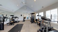Located at 1400 Laurel Springs Dr in Durham, NC, this single-floor fitness center is designed to provide a welcoming and functional workout environment. The room is defined by its striking vaulted ceiling, which lends a sense of openness and airiness to the space. Dual ceiling fans circulate air throughout, while recessed lighting ensures the area is well-lit at all times. Large windows along the walls invite generous natural light, further enhancing the bright and energizing atmosphere. The fitness center is equipped with a comprehensive selection of exercise equipment, including treadmills, a Landice cardio machine, a multi-station cable and weight machine, a workout bench, and yoga/exercise mats for floor-based training. The light-toned, textured flooring provides a clean and durable surface suited for a variety of workout activities. A classic round wall clock is mounted near the entrance for easy time-keeping during workouts. French glass doors at the rear of the room offer a welcoming entry point and allow additional natural light to filter in. The overall ambiance is clean, spacious, and motivating — well-suited for a range of fitness routines from strength training to cardio and stretching.