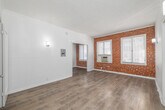 Located at 701 S Gramercy Dr in Los Angeles, CA, this single-floor unit offers a thoughtfully laid-out living space with a living room and two bathrooms. The living room immediately draws the eye with its striking exposed red brick accent wall, which adds warmth and character to the otherwise crisp, white-painted interior. Light hardwood flooring runs throughout, enhancing the bright and airy feel of the space. Multiple windows with blinds allow natural light to filter in, complementing the textured white ceilings. An in-wall air conditioning unit provides climate control. The layout opens through a doorway into an adjoining area, creating a natural flow between spaces. One of the bathrooms features classic black-and-white octagonal tile flooring, a white bathtub with an overhead shower, navy tile accents, and a window for natural ventilation. The second bathroom area is highlighted by a built-in floating vanity with multiple drawers, a vertical mirror with overhead lighting, and an adjacent storage unit, offering practical functionality with a clean aesthetic. Together, the exposed brick, hardwood floors, and white walls create a balanced blend of industrial charm and modern simplicity throughout this Los Angeles residence.