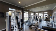 This fitness center offers a thoughtfully designed workout environment built to support a full range of exercise routines. The space is anchored by a robust collection of strength-training equipment, including cable machines, multi-function weight stations, and free weights such as barbells and weight plates, providing options for users of all fitness levels. Cardio equipment is also present, rounding out a well-balanced facility. The flooring features durable, dark rubber matting suited for heavy use and easy maintenance. Large, multi-pane windows line the walls, flooding the space with natural light and offering views of the surrounding exterior. Full-length mirrors are mounted along one wall, enhancing the sense of openness and allowing users to monitor their form during workouts. The ceiling showcases elegant crown molding and recessed lighting, lending a polished, refined aesthetic that sets this facility apart from a standard gym environment. The overall color palette of deep taupe walls and dark flooring creates a focused, professional ambiance. The single-floor layout ensures easy accessibility and a streamlined flow throughout the space, making it both functional and visually cohesive.