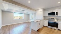 Located at 925 Mix Ave in Hamden, Connecticut, this thoughtfully designed unit offers a clean, modern aesthetic across a single floor. The open-concept layout seamlessly connects the kitchen and living room, creating a bright and functional living space. Warm-toned hardwood-style flooring runs throughout, complementing the neutral gray walls and crisp white trim.

The kitchen is a standout feature, outfitted with white shaker-style cabinetry, granite-look countertops, and a peninsula island that doubles as additional prep and dining space. Stainless steel appliances — including a range, over-the-range microwave, and refrigerator — complete the well-appointed setup. Recessed lighting and a ceiling-mounted HVAC unit add to the modern, polished feel.

The living area benefits from large windows that invite natural light and offer views of the surrounding neighborhood, enhancing the open and airy ambiance of the space. Additional rooms include a bright, neutral-toned room with a window framing outdoor views — ideal as a bedroom or flex space — and a clean, all-white bathroom featuring a full tub/shower combination with built-in shelving. A hallway connects the rooms, also finished with the same consistent flooring and white millwork, tying the entire unit together cohesively.