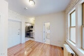 This compact single-floor unit showcases a blend of classic architectural character and modern updates. The main living area is defined by white-painted exposed brick walls that lend a distinctive urban aesthetic, complemented by warm hardwood flooring that runs throughout. Natural light filters in through double-pane windows, illuminating the space while a classic cast-iron radiator provides heating. A built-in shelving nook adds both charm and practical storage, and a closet with a bifold door offers additional organization.

The kitchen is efficiently designed, featuring stainless steel appliances including a gas range, refrigerator, dishwasher, and microwave, paired with white cabinetry and clean countertops. Marble-look tile flooring distinguishes the kitchen area from the rest of the unit. The entry door opens directly into the kitchen, creating a functional and straightforward layout.

The bathroom is crisp and well-appointed, with white subway tile lining the bathtub surround, a tub-shower combination with chrome fixtures, and a clean vanity. The overall ambiance of the unit is bright and airy, with the white-painted brick tying each area together into a cohesive, light-filled space that balances historic building character with contemporary finishes.