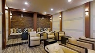 This elegant single-floor lounge exudes a refined ambiance through its thoughtful combination of natural and contemporary design elements. The most striking feature is the full-height stacked stone accent wall, rendered in warm earth tones of amber, rust, and brown, which serves as a dramatic backdrop to the seating area. Richly stained wooden columns frame the stone wall, adding structural character and warmth throughout the space. The seating arrangement is both generous and well-curated, featuring plush cream and beige upholstered armchairs and sofas accented with bold black-and-white geometric throw pillows, all complemented by sleek dark-framed side tables. Recessed ceiling lighting and softly glowing wall sconces cast a warm, layered light throughout the room, enhancing the inviting atmosphere. The flooring features a subtly patterned carpet in neutral tones that ties the color palette together cohesively. On the opposing wall, a clean, paneled white surface with horizontal detailing provides a calm contrast to the textured stone, balancing the overall aesthetic. The space is designed to encourage comfortable gathering, with furniture groupings that promote both intimate conversation and open relaxation in an upscale, lounge-style setting.