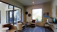 This single-story space is designed to create a warm and inviting atmosphere for visitors from the moment they arrive. The interior is defined by neutral, warm-toned walls that lend a calm and professional aesthetic throughout. The waiting area serves as the focal point of the space, thoughtfully appointed with contemporary leather and upholstered seating arranged to encourage comfort and ease. Small accent tables are positioned conveniently between chairs, adding both functionality and a refined touch to the layout. Dark hardwood-style flooring grounds the room with a rich contrast against the lighter walls and furnishings. Large, black-framed windows allow generous amounts of natural light to flood the interior while offering pleasant views of the surrounding greenery and landscaping outside. Decorative circular wall art adds a subtle artistic element to the space without overwhelming its clean, understated design. Recessed ceiling lighting ensures the area remains well-lit and welcoming regardless of the time of day. Glass entry doors with a sleek dark frame provide a modern, transparent boundary between the interior and the exterior, further enhancing the sense of openness. Overall, the space balances professionalism with approachability, making it well-suited for a client-facing or public-facing commercial use.