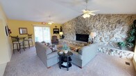 This single-floor space makes an immediate impression with its commanding floor-to-ceiling stone fireplace wall, which serves as the defining architectural centerpiece of the main living area. The natural stonework spans the full height of the vaulted ceiling, creating a striking focal point that lends the space a rustic yet refined character. The vaulted ceiling itself adds a generous sense of openness and volume, further enhanced by multiple ceiling fans that provide both comfort and visual interest overhead.

The living area is generously sized, accommodating a comfortable seating arrangement with plush sofas, accent chairs, and a decorative area rug positioned in front of the fireplace hearth. Warm yellow walls complement the natural tones of the stonework, contributing to an inviting and cheerful ambiance. A sliding glass door and several windows dressed with decorative valances allow natural light to filter in, offering views of the surrounding outdoor greenery.

The kitchen features cream-toned cabinetry, a white tile backsplash, and a full suite of appliances including a refrigerator, range, dishwasher, and microwave. A double sink and ample counter space support practical everyday use. Consistent yellow wall tones and a decorative wallpaper border tie the kitchen's aesthetic to the rest of the space, creating a cohesive and homey feel throughout.