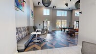 Located at 2000 Brandt Dr in Cranberry Township, PA, this single-floor community lounge showcases a contemporary design with a distinctly open, airy atmosphere. The space is immediately defined by its dramatic double-height ceilings, which allow natural light to pour in through large upper-level windows and floor-to-ceiling glass panels along the exterior wall, offering views of the outdoors.

One of the most striking architectural features is the bold chevron-patterned flooring, composed of contrasting dark and light wood tones, which creates a dynamic visual foundation throughout the space. The lounge area near the entrance features built-in banquette seating upholstered in patterned fabric, complemented by modern swivel chairs and bistro-style tables—ideal for casual gatherings or individual use. Colorful abstract artwork adorns the neutral gray walls, adding vibrant character.

Toward the rear, a recreation area with a pool table provides an engaging social focal point, surrounded by additional lounge seating. A well-appointed kitchenette lines one wall, equipped with stainless steel appliances including a full-size refrigerator, microwave, and white cabinetry with dark countertops. A wall-mounted flat-screen display and integrated power outlets throughout enhance functionality, making this a versatile, amenity-rich communal space.