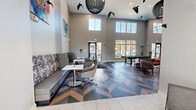 Located at 2000 Brandt Dr in Cranberry Township, PA, this single-floor community lounge showcases a contemporary design with a distinctly open, airy atmosphere. The space is immediately defined by its dramatic double-height ceilings, which allow natural light to pour in through large upper-level windows and floor-to-ceiling glass panels along the exterior wall, offering views of the outdoors.

One of the most striking architectural features is the bold chevron-patterned flooring, composed of contrasting dark and light wood tones, which creates a dynamic visual foundation throughout the space. The lounge area near the entrance features built-in banquette seating upholstered in patterned fabric, complemented by modern swivel chairs and bistro-style tables—ideal for casual gatherings or individual use. Colorful abstract artwork adorns the neutral gray walls, adding vibrant character.

Toward the rear, a recreation area with a pool table provides an engaging social focal point, surrounded by additional lounge seating. A well-appointed kitchenette lines one wall, equipped with stainless steel appliances including a full-size refrigerator, microwave, and white cabinetry with dark countertops. A wall-mounted flat-screen display and integrated power outlets throughout enhance functionality, making this a versatile, amenity-rich communal space.