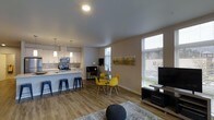 This single-floor space showcases a thoughtfully designed open-concept layout that seamlessly connects the kitchen, dining, and living areas into one cohesive and functional environment. The modern kitchen is outfitted with stainless steel appliances, white cabinetry, a tile backsplash, and a peninsula island with bar seating — ideal for casual dining or entertaining. Pendant lighting above the island adds a stylish focal point, complemented by a track lighting system overhead. The dining area features a glass-top table with vibrant yellow chairs, contributing a pop of color against the neutral-toned walls. Large windows fitted with horizontal blinds invite generous natural light throughout the space, creating a bright and airy atmosphere. Warm-toned hardwood-style flooring runs throughout, tying the open space together with a clean, contemporary aesthetic. The living area is defined by a patterned area rug and provides a comfortable setting adjacent to the main living zones. A dedicated workspace with a compact desk is thoughtfully tucked near the windows, offering a functional area for productivity. The overall ambiance is modern, welcoming, and efficiently arranged.