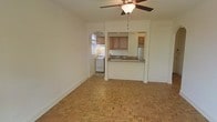 This single-floor space combines classic design elements with practical functionality. The living area is immediately welcoming, featuring warm parquet wood flooring laid in a distinctive geometric pattern that adds character and visual interest underfoot. A ceiling fan with a light fixture is centrally positioned, ensuring comfortable airflow throughout the space.

An elegant arched passageway connects the living area to the kitchen, lending an architectural charm that sets this space apart. The kitchen is thoughtfully designed with a convenient pass-through bar counter topped with granite, making it ideal for casual dining or entertaining. Warm-toned wooden cabinetry provides ample storage, while a gas range and a full-sized refrigerator complete the kitchen's practical amenities. Natural light filters in through a window adjacent to the range, brightening the cooking space.

The overall aesthetic is clean and neutral, with white walls throughout that provide a versatile backdrop for a variety of décor styles. The combination of the arched entryway, parquet flooring, and granite countertops gives the space a timeless quality, blending traditional architectural details with functional, everyday living essentials.