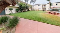 This ground-floor unit, numbered 106, is situated within a multi-building residential complex that showcases a well-maintained outdoor environment and a range of shared amenities. The exterior of the complex features stucco-finished buildings with a neutral beige palette, complemented by dark-trimmed balconies and staircases serving upper-level units.

The unit's entrance is framed by a distinctive dark green door, flanked by a window with security bars and surrounded by small planted shrubs and decorative items, lending the entry a personalized touch. A red-toned concrete pathway leads to the front door, blending with the broader hardscape that winds throughout the complex.

The shared outdoor spaces are a notable highlight of this property. A spacious, well-kept lawn area provides open green space, bordered by mature trees including towering palms and weeping varieties that lend a California ambiance. A charming courtyard garden features an eclectic mix of potted plants, ornamental grasses, and established trees, creating a lush, layered landscape.

The complex also includes a fenced communal swimming pool, complete with poolside seating and umbrella shading, surrounded by manicured plantings and additional hardscaped areas — ideal for outdoor relaxation.