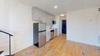 Located at 2009 Greene St in Columbia, SC's vibrant 29205 corridor, this two-floor loft-style space masterfully combines historic industrial character with contemporary finishes. The main level immediately impresses with its original hardwood floors bearing the rich patina of age, an exposed brick accent wall with weathered texture, and a bold black spiral staircase that serves as both a functional element and a visual centerpiece. A large multi-pane window floods the space with natural light while framing views of the surrounding neighborhood. The kitchen is efficiently designed with gray cabinetry extending to the ceiling, stainless steel appliances including a full-size refrigerator, built-in microwave, and dishwasher, complemented by clean white countertops and a sink. A full bathroom on the main level features large-format tile flooring, a dark wood vanity with a wide mirror, and a tiled tub-shower combination with decorative mosaic accent tile. A convenient half-bath with a pedestal sink and round mirror provides additional functionality. The upper level offers a carpeted bedroom retreat with soaring ceilings, exposed structural beams, and an en suite bathroom, creating a private and comfortable sanctuary above the open living areas below.