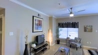 This single-floor space offers a thoughtfully designed living area that combines comfort and style. The living room serves as the centerpiece, featuring elegant crown molding that frames the ceiling and adds a refined architectural detail to the room. A ceiling fan with a built-in light fixture enhances air circulation while contributing to the room's aesthetic appeal. Natural light filters in through a large window dressed with patterned curtains and blinds, creating a bright and welcoming atmosphere. The neutral-toned walls provide a versatile backdrop that complements the room's furnishings and decor. The carpeted flooring adds warmth and comfort underfoot. A dedicated media area accommodates a flat-screen television on a sleek media console, while a cozy reading nook near the window features a patterned armchair and a small desk, ideal for relaxing or working. Decorative touches, including framed wall art, indoor plants, and accent vases, bring personality and life to the space. The open layout allows for easy furniture arrangement and a natural flow throughout the room, making it both functional and inviting for everyday living.