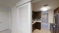 This single-floor space showcases a thoughtfully designed kitchen area visible through a sleek, white louvered sliding door — a standout architectural detail that adds both visual interest and practical flexibility to the layout. The sliding door features clean horizontal slats and glides along an overhead track, allowing the kitchen to be opened to or separated from the adjacent space as needed.

The kitchen itself is outfitted with rich, dark espresso-toned cabinetry featuring brushed silver hardware, offering ample upper and lower storage. A light-colored countertop provides a bright contrast against the darker cabinet tones, while a mosaic tile backsplash in a mix of neutral and earthy tones adds texture and visual depth to the space.

Overhead, a flush-mounted circular ceiling light provides even illumination, complemented by track lighting that offers directed task lighting throughout the area. The light-toned tile flooring ties the space together, reflecting light and contributing to an open, airy feel. Overall, the space balances modern design elements with functional details, creating a practical yet visually cohesive interior.