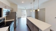 This well-appointed single-floor residence offers a thoughtfully designed layout that balances modern aesthetics with everyday functionality. The open-concept kitchen serves as the centerpiece of the living area, featuring warm wood-toned cabinetry, a classic subway tile backsplash, stainless steel appliances including a range, microwave, dishwasher, and refrigerator, and striking quartz countertops. A large kitchen island with pendant lighting above provides additional prep space and a natural gathering point, while an open view toward the living area and sliding glass door leads to a private balcony beyond. The living area benefits from dark hardwood-style flooring and a ceiling fan, creating a comfortable and cohesive space. The bedroom is bright and airy, filled with natural light from a wide window fitted with blinds, complemented by soft carpet underfoot and a ceiling fan overhead. The full bathroom is finished with wood-look plank flooring, a dual-sink vanity with clean white countertops, and a soaking tub and shower enclosed in crisp white subway tile. A generously sized walk-in closet features wall-to-wall wire shelving and drawer units, offering extensive organization and storage capacity. Throughout the residence, a neutral palette and contemporary finishes create a clean, modern ambiance.