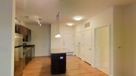 This single-floor unit offers a well-organized layout that maximizes its available space through an open-concept design. The kitchen is a standout feature, outfitted with dark espresso cabinetry, a stainless steel range, and a central island topped with a light stone-style countertop. A pendant light hangs directly above the island, complementing the track lighting nearby and the flush-mount ceiling fixtures that provide ample illumination throughout. The kitchen flows naturally into the main living area, which is finished with warm hardwood-style flooring that extends throughout much of the unit. The living space benefits from a window that brings in natural light, contributing to an open and airy atmosphere. Interior doors with paneled detailing lead to additional areas, including what appears to be a bathroom complete with standard fixtures. The neutral white walls throughout the unit provide a clean, versatile backdrop. The overall layout is practical and efficient, with clearly defined zones for cooking, living, and personal use, making the space functional for everyday living.