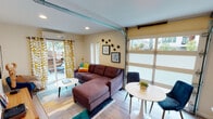 Located at 2120 N Garrett Ave in Dallas, TX, this single-story studio offers a thoughtfully designed open-concept layout that maximizes its compact footprint. The living area serves as the centerpiece of the space, defined by a striking retractable glass garage door that floods the interior with natural light and provides a seamless visual connection to the outdoors. Light wood-style flooring runs throughout, complementing the warm, neutral wall tones and lending a cohesive, contemporary feel.

The living area comfortably accommodates a sectional sofa and a small dining table with seating, making efficient use of the available space. A wall-mounted mini-split system provides climate control, while recessed lighting adds a modern touch overhead. Sliding glass doors along one wall open to a private outdoor deck, further enhancing the sense of openness.

The kitchen is efficiently laid out in a galley configuration, featuring dark espresso cabinetry, white countertops, stainless steel appliances including a refrigerator, electric range, and microwave, and a single-basin sink. A pass-through breakfast bar with bar stools connects the kitchen to the main living area, promoting an open, social atmosphere.

The sleeping area is delineated by a curtained partition, offering a degree of privacy. The bathroom includes a full tub-shower combination finished with large-format gray tile, wood-look flooring, and clean, simple fixtures.