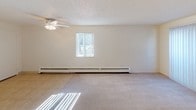 This single-floor unit offers a practical and straightforward layout designed for efficient use of space. The main living area is carpeted and bathed in natural light through a large sliding glass door fitted with vertical blinds, which opens to an outdoor view. A ceiling fan with a light fixture is centrally mounted on the textured ceiling, complementing the baseboard heating that runs along the exterior wall. A wall-mounted air conditioning unit provides additional climate control.

The kitchen is positioned along one wall and features light wood cabinetry, a laminate countertop, a single-basin sink, a white electric range, and a dishwasher. Tile flooring and overhead fluorescent lighting give the kitchen a clean, utilitarian appearance. An electrical panel is conveniently accessible near the entryway.

The bathroom area includes a vanity with a stone-toned laminate countertop, a white basin sink, light wood cabinetry below, and a large mirror with overhead Hollywood-style lighting. Adjacent to the bathroom is a walk-in closet with a single hanging rod and carpeted flooring, providing functional storage. Overall, the unit presents a neutral, move-in-ready aesthetic with practical amenities throughout.