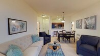 This single-floor residence offers a thoughtfully designed open-concept layout that seamlessly connects the living, dining, and kitchen areas into one cohesive and functional space. The living room features plush carpeted flooring, a tufted sofa with teal accent pillows, and a cushioned armchair, creating a welcoming and relaxed atmosphere. A patterned area rug anchors the seating arrangement around a central coffee table. Adjacent to the living area, the dining space accommodates a full-sized table with seating for multiple guests, ideal for everyday meals and entertaining. The kitchen is positioned at the far end of the open layout, featuring rich dark wood cabinetry, black appliances, a raised breakfast bar with bar stool seating, and a stylish pendant light overhead that adds a touch of warmth. Recessed lighting throughout ensures the space remains bright and inviting. A dedicated laundry area is visible through a doorway, adding everyday convenience. The neutral color palette of soft whites and warm beiges throughout the space contributes to an airy, cohesive aesthetic. The open floor plan maximizes the sense of space, making this residence both practical and comfortable for daily living.