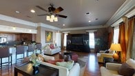 This single-floor residence offers a thoughtfully designed living area that balances comfort and style. The main living room serves as the centerpiece of the home, featuring warm hardwood flooring that extends throughout the open layout. Crown molding lines the upper perimeter of the walls, adding a refined architectural detail that complements the overall aesthetic.

A ceiling fan with a built-in light fixture is centrally mounted, providing both functionality and visual appeal. Recessed lighting further illuminates the space, creating a well-lit and inviting atmosphere. The room is anchored by a large, dark wood entertainment unit housing a flat-screen television, flanked by decorative columns that add a sense of structure and sophistication.

Seating is generously arranged, with plush sofas and accent chairs surrounding a glass-top coffee table set atop an area rug, defining the conversational zone. Decorative throw pillows in warm tones add pops of color. A framed piece of artwork adorns one wall, while an indoor plant introduces a natural, organic element. Natural light filters in through curtained windows, enhancing the warm and welcoming ambiance throughout the space.