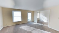 This single-floor space offers a clean, functional layout designed to maximize comfort and usability. The main living area is generously proportioned, finished with wall-to-wall neutral-toned carpet that adds warmth underfoot. Soft yellow walls create a welcoming ambiance throughout, complementing the bright, airy feel of the interior.

Natural light is a defining feature of the living space, with multiple windows fitted with horizontal blinds lining the exterior wall, along with a glass-paneled rear door that invites an abundance of daylight into the room. The white smooth ceilings are clean and bright, further enhancing the open, uncluttered feel of the space.

An open passage leads to an adjacent room or hallway, suggesting a well-connected floor plan that encourages easy movement between areas. The staircase visible in the background hints at additional vertical access or a split-level element within the layout.

Floor-mounted heating vents are positioned along the base of the walls, ensuring efficient climate control throughout the space. The overall aesthetic is simple and neutral, making it highly versatile and easy to personalize. This space is well-suited for those seeking a bright, open, and move-in-ready environment.