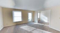 This single-floor space offers a clean, functional layout designed to maximize comfort and usability. The main living area is generously proportioned, finished with wall-to-wall neutral-toned carpet that adds warmth underfoot. Soft yellow walls create a welcoming ambiance throughout, complementing the bright, airy feel of the interior.

Natural light is a defining feature of the living space, with multiple windows fitted with horizontal blinds lining the exterior wall, along with a glass-paneled rear door that invites an abundance of daylight into the room. The white smooth ceilings are clean and bright, further enhancing the open, uncluttered feel of the space.

An open passage leads to an adjacent room or hallway, suggesting a well-connected floor plan that encourages easy movement between areas. The staircase visible in the background hints at additional vertical access or a split-level element within the layout.

Floor-mounted heating vents are positioned along the base of the walls, ensuring efficient climate control throughout the space. The overall aesthetic is simple and neutral, making it highly versatile and easy to personalize. This space is well-suited for those seeking a bright, open, and move-in-ready environment.