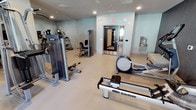 This single-floor fitness center offers a clean, functional environment designed to support a full range of workout routines. The space features light-toned flooring and soft blue-green walls that create a calm, focused atmosphere throughout. Recessed lighting illuminates the room evenly, ensuring a bright and welcoming workout environment.

The facility is outfitted with a thoughtfully curated selection of commercial-grade equipment. A WaterRower rowing machine takes a prominent position on the floor, offering a low-impact, full-body cardio option. Nearby, an elliptical trainer provides additional cardiovascular options alongside a Precor-branded upright cycle. For strength training, a cable machine and a seated weight station are available, catering to a variety of muscle groups and fitness levels.

A wall-mounted mirror on one side of the room enhances the sense of space while allowing users to monitor their form during exercise. A wall-mounted water fountain is conveniently located near the exit, keeping hydration accessible at all times. The room is accessed through a set of glass-paneled doors, contributing to a polished, professional aesthetic. Overall, this fitness center is a practical and well-appointed space suited for daily use.