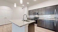 This thoughtfully designed one-bedroom, one-bathroom unit offers a modern and cohesive aesthetic across its single floor. The open-concept kitchen and living area serves as the heart of the space, where light wood-tone flooring flows seamlessly underfoot. The kitchen is a standout feature, showcasing dark espresso cabinetry with sleek hardware, granite countertops, a white subway tile backsplash, and a full suite of stainless steel appliances including a range, microwave, and refrigerator. A granite-topped kitchen island with an undermount sink and pendant lighting above adds both functionality and style, creating a natural gathering point that connects the kitchen to the living area.

A hallway leads to the generously sized primary bedroom, which is carpeted for comfort and features a dedicated closet outfitted with wire shelving and a laundry access panel. The bathroom is clean and bright, offering light wood-tone flooring, dark cabinetry with granite counters, wire shelving for storage, and a full soaking tub with shower. Neutral wall tones and recessed lighting throughout contribute to a bright, cohesive ambiance across the entire unit.