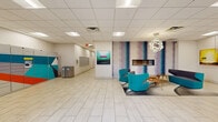 Located at 115 Second Ave S in downtown Minneapolis, this lobby offers a well-appointed, single-floor space designed to serve as an efficient and inviting gateway. The entrance features large glass doors flanked by structural columns, allowing natural light to filter in and providing views of the surrounding streetscape. Light-toned tile flooring runs throughout, creating a clean, cohesive aesthetic.

A central lounge area anchors the space, furnished with vibrant teal swivel chairs and a mid-century style wood coffee table, all set atop a layered area rug. A linear gas fireplace is set into a bold, artistically muraled accent wall in purple and teal tones, while a sculptural orb pendant light hangs overhead, adding a contemporary focal point. Curated artwork adorns the walls throughout, contributing to an engaging visual environment.

A glass-enclosed office or leasing suite provides a transparent, professional workspace visible from the main lobby. The package management area houses a Luxer One smart locker system, offering a secure and convenient parcel solution. Mailboxes and additional utility elements are also present. The drop ceiling with recessed and panel lighting ensures consistent, comfortable illumination throughout. This lobby is thoughtfully designed to balance functionality with modern style.