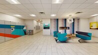 Located at 115 Second Ave S in downtown Minneapolis, this lobby offers a well-appointed, single-floor space designed to serve as an efficient and inviting gateway. The entrance features large glass doors flanked by structural columns, allowing natural light to filter in and providing views of the surrounding streetscape. Light-toned tile flooring runs throughout, creating a clean, cohesive aesthetic.

A central lounge area anchors the space, furnished with vibrant teal swivel chairs and a mid-century style wood coffee table, all set atop a layered area rug. A linear gas fireplace is set into a bold, artistically muraled accent wall in purple and teal tones, while a sculptural orb pendant light hangs overhead, adding a contemporary focal point. Curated artwork adorns the walls throughout, contributing to an engaging visual environment.

A glass-enclosed office or leasing suite provides a transparent, professional workspace visible from the main lobby. The package management area houses a Luxer One smart locker system, offering a secure and convenient parcel solution. Mailboxes and additional utility elements are also present. The drop ceiling with recessed and panel lighting ensures consistent, comfortable illumination throughout. This lobby is thoughtfully designed to balance functionality with modern style.