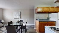 This single-floor unit offers a functional and comfortable layout with distinct living spaces thoughtfully arranged for everyday living. The living room is anchored by a large window fitted with blinds, allowing natural light to fill the room, which features dark wood-look flooring, neutral gray walls, and a baseboard heating unit. A glass-top coffee table and a contemporary sofa with mixed accent pillows complete the space. Adjacent to the living room, the dining area accommodates a glass-top dining table with seating for four, set against a soft neutral wall accented with decorative artwork, and flows seamlessly into the kitchen. The galley-style kitchen is outfitted with warm oak cabinetry, granite-look countertops, a tile backsplash, a gas range, a refrigerator, and a stainless steel sink — all finished with light ceramic tile flooring. The bedroom is a serene retreat featuring the same wood-look flooring, soft neutral walls, multiple windows, a wall-mounted air conditioning unit, and a baseboard heater. The full bathroom includes a vanity with a mirrored medicine cabinet, a toilet, and a combination tub-shower, complemented by classic mosaic tile flooring. Built-in shelving adjacent to the bathroom provides convenient additional storage.