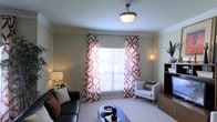 This single-floor living space presents a thoughtfully designed interior centered around a comfortable and stylish living room. The room features neutral beige walls accented by crisp white crown molding that runs along the ceiling perimeter, adding a classic architectural touch. A flush-mount ceiling light fixture with a brushed nickel finish serves as the central lighting source, complemented by a tall floor lamp and a table lamp that provide warm ambient lighting throughout the space.

A large window, dressed with bold rust-and-white patterned curtains featuring a geometric floral motif, serves as the focal point of the room, allowing natural light to filter in through white horizontal blinds. The soft, light-colored carpeting underfoot adds warmth and comfort to the space.

Furnishings include a dark leather sofa adorned with decorative pillows and a crisp white accent chair with coordinating cushions, arranged around a rectangular coffee table that anchors the seating area. A bookcase or media unit is partially visible along one wall. The overall ambiance is warm, coordinated, and inviting, blending classic architectural details with contemporary décor elements.