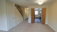 This two-story residence offers a functional and straightforward layout across its two floors. The main level features a spacious carpeted living area with neutral-toned walls and flush ceiling lighting, creating a clean and welcoming atmosphere. A staircase with decorative wrought-iron balusters rises along one wall, providing an understated architectural detail that adds character to the space.

Adjacent to the living area, the kitchen is accessible through an open doorway framed by warm wood-toned cabinetry panels. The kitchen is equipped with dark appliances, including a refrigerator and range, set against wood-finish cabinetry that lines the upper walls. A window above the counter allows natural light to brighten the cooking space. The kitchen flooring contrasts with the carpeted living area, featuring a darker hard-surface material that defines the two zones.

A hallway visible from the main living area leads to additional rooms on the ground floor, with multiple doors suggesting closet or utility spaces. The overall ambiance of the home is practical and comfortable, with a neutral palette throughout that provides a versatile backdrop for a variety of interior styles.