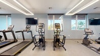 This fitness center offers a comprehensive and thoughtfully arranged workout environment spread across a single floor. The space is designed with a clean, modern aesthetic, featuring light blue walls, neutral carpet flooring, and a combination of recessed and linear LED lighting that ensures the entire area is evenly and brightly illuminated.

The cardio zone is well-appointed with multiple treadmills, elliptical trainers, stair climbers, and a rowing machine, positioned near large windows fitted with horizontal blinds. These windows allow natural light to filter in and provide views of the surrounding outdoor environment, creating an energizing atmosphere for exercise.

The strength training area features a dedicated free weights section with a dumbbell rack, adjustable bench, and cable machines, all positioned in front of a full-length mirror wall that visually expands the space and aids in form correction. Additional cable and resistance machines are organized throughout the floor, offering a wide range of training options.

A warm-toned wooden shelving unit provides useful storage near the entrance. Wall-mounted televisions are positioned throughout for entertainment during workouts. The space is accessed through sturdy double doors and maintains a spacious, open layout that accommodates multiple users comfortably.