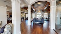 This single-story clubhouse showcases an impressive open-concept layout defined by rich dark hardwood flooring, soaring coffered ceilings with exposed wood beams, and elegant white structural columns framed by sweeping archways. The expansive great room serves as the centerpiece, anchored by a whitewashed brick fireplace with a wall-mounted television and built-in shelving, complemented by stylish seating arrangements and a statement chandelier overhead. Large arched windows and glass-paneled doors flood the space with natural light while offering views of the surrounding outdoor area. A dedicated dining nook features an octagonal wood table beneath tall grid-paned windows with floor-length drapes, positioned adjacent to a kitchenette with modern appliances and white cabinetry. The game room provides a lively recreational retreat, complete with a billiards-style table, wall-mounted television, and pendant lighting. A quieter lounge area offers a more intimate setting, finished with shiplap accent walls, carpeted floors, wall sconces, and ample natural light through slatted blinds. Throughout, the space balances warmth and sophistication, blending transitional design elements with inviting, community-focused amenities ideal for both relaxation and social gathering.