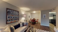 This single-floor residence at Breckinridge Square offers a thoughtfully designed open layout that seamlessly connects the living and dining areas. The living space is anchored by a comfortable sofa accented with decorative pillows and complemented by a bold, framed abstract artwork that adds a pop of color to the neutral gray walls. A stylish table lamp provides warm ambient lighting, enhancing the inviting atmosphere of the room.

The rich, dark hardwood-style flooring flows continuously throughout, creating a cohesive and polished look across both the living and dining areas. The dining space is defined by a round table paired with vibrant orange chairs, adding a striking contrast to the otherwise understated color palette. A wainscoting detail on the dining room wall introduces a refined architectural touch.

The textured ceiling adds subtle visual interest overhead, while clean white baseboards frame the space neatly. The open sightlines between rooms create a sense of spaciousness and connectivity, making the layout feel functional and well-proportioned. Overall, the space strikes a balance between contemporary style and comfortable everyday living.