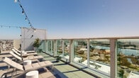 Located at 255 W 5th St in San Pedro, California, this rooftop terrace offers an exceptional outdoor experience defined by sweeping panoramic views of the Los Angeles harbor and surrounding cityscape. Floor-to-ceiling glass railings line the perimeter, ensuring unobstructed sightlines across the waterfront while maintaining an open, airy atmosphere under the Southern California sky.

The terrace is thoughtfully organized into several distinct functional zones. A dedicated lounge area features cushioned chaise lounges and low-profile seating, ideal for relaxing and taking in the harbor views. Multiple conversation seating clusters, furnished with wicker chairs and side tables, are distributed across the tiled deck, providing intimate spaces for gatherings. Overhead string lights span the entire terrace, adding a warm, ambient glow for evening use.

A well-appointed outdoor kitchen and BBQ station anchors one side of the roof, equipped with multiple grills set against a bold geometric patterned tile accent wall. Adjacent dining tables and chairs offer ample seating for al fresco meals. Potted palms and planters introduce a touch of greenery throughout the space. The large-format tile flooring in a neutral palette ties the entire terrace together, creating a cohesive and polished aesthetic suited for both social events and everyday relaxation.
