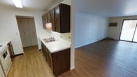 This single-floor space is designed with a practical, open layout that connects the kitchen and living area seamlessly. The kitchen features dark wood cabinetry extending from floor to ceiling, providing ample storage. A white countertop runs along the length of the kitchen, housing a stainless steel double-basin sink positioned beneath under-cabinet lighting. The lower cabinetry mirrors the upper units in dark wood tones, maintaining a cohesive aesthetic throughout the space. Light wood-tone laminate flooring runs continuously through both the kitchen and the adjoining living area, creating visual unity across the open floor plan. The living area is notably open and bright, with white walls and clean white baseboards that lend a fresh, neutral backdrop. A baseboard heater is visible along the far wall of the living area, indicating an efficient heating system. The transition between the kitchen and living space is defined by a partial wall, adding subtle architectural separation while maintaining the open feel. Overall, the space presents a clean, functional design well-suited for everyday living.