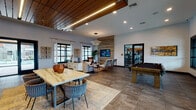 This single-floor community clubhouse is thoughtfully designed to blend modern aesthetics with warm, inviting materials. The main gathering area is anchored by a striking slatted wood accent wall that frames a wall-mounted television, creating a natural focal point for the lounge seating arrangement. Comfortable sofas, accent chairs, and layered area rugs define the relaxed social zone, while large black-framed windows allow abundant natural light to fill the space. A wood-paneled dropped ceiling with integrated linear LED lighting adds architectural depth and warmth overhead, contrasting beautifully with the large-format stone tile flooring throughout. The bar area features a dramatic, veined stone waterfall island with wire-frame barstools, set against a bold patterned accent wall and dark cabinetry housing a coffee station and flat-screen display. A full-size pool table anchors the game zone, conveniently adjacent to glass-paned doors leading into a well-equipped fitness center. The entry lobby offers a welcoming reception desk with oversized windows overlooking the exterior. Curated artwork, lush greenery, and eclectic furnishings complete the cohesive, upscale-casual atmosphere throughout this amenity-rich space.