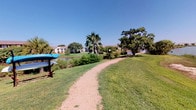 This outdoor recreational area offers a well-maintained, park-like environment designed for leisure and active use. A winding gravel path serves as the central feature, meandering through expansive, manicured green lawns beneath a clear, open sky. The grounds are generously shaded by a mix of mature oak trees and tall palm trees, lending the space a distinctive character that balances tropical and traditional landscaping aesthetics.

A notable waterfront element runs along one side of the path, where a calm body of water bordered by natural marsh grasses provides scenic views and a tranquil atmosphere. A sandy gravel area near the water's edge appears designated for watercraft access, with kayak storage racks visible nearby, indicating direct recreational water access for residents.

Path lighting posts are positioned throughout the grounds, suggesting the area is accessible and usable during evening hours. Lush, well-trimmed shrubs and layered plantings line the buildings' exteriors, creating a polished and welcoming perimeter. Concrete sidewalks wind alongside building facades, connecting various areas of the community seamlessly.

Overall, the recreational area presents a vibrant, active outdoor environment with a strong connection to nature, offering residents open green space, waterfront access, and shaded walkways throughout the community.