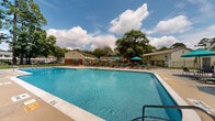 Located at 301 Village Dr in Jacksonville, North Carolina, this outdoor pool facility offers a well-appointed recreational space designed for relaxation and leisure. The centerpiece is a generously sized swimming pool with clear, blue water, framed by a spacious concrete sundeck that provides ample room for sunbathing and socializing. The deck is outfitted with numerous lounge chairs and teal-accented umbrellas, offering both sun and shade options throughout the day. A dedicated seating area near the pool house features additional chairs and tables, ideal for casual gatherings. The pool area is fully enclosed by a sturdy black iron fence, ensuring safety and privacy. A utility building adjacent to the pool house houses the mechanical equipment supporting the facility. Mature trees — including tall pines and broad hardwoods — surround the perimeter, providing a natural backdrop and pleasant shade. Well-maintained green lawns border the fenced enclosure, enhancing the overall aesthetic. The residential buildings of the community are visible in the background, reinforcing the community-oriented nature of this amenity. Under open Carolina skies, this pool area serves as an inviting outdoor retreat for residents of the surrounding community.