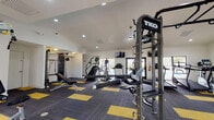 This fitness center is organized across a single level and designed to support a comprehensive range of workout styles. The main workout area features soaring vaulted ceilings with crown molding, recessed lighting, and a large ceiling fan that keeps the space well-ventilated. The flooring throughout consists of a distinctive charcoal and yellow modular carpet tile pattern that adds a bold, athletic aesthetic. Large windows and glass double doors invite natural light and provide views of an adjacent outdoor pool area.

The main floor is outfitted with TKO multi-functional training rigs, including monkey bars, pull-up stations, and a hanging heavy punching bag — ideal for functional and combat fitness training. Cardio equipment includes Matrix treadmills, elliptical machines, and stair climbers, while strength training is supported by cable machines, a Hoist weight machine, a rowing machine, a flat bench, and a dumbbell rack.

A separate, quieter room with teal accent walls and windows overlooking the pool serves as a dedicated cycling studio, housing multiple red stationary spin bikes and a storage box. An additional room with matching teal accents and ample natural light provides space for a rowing machine, a weight machine, and a dumbbell rack. Wall-mounted mirrors throughout the facility enhance the sense of space and allow for form monitoring during exercise.
