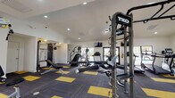 This fitness center is organized across a single level and designed to support a comprehensive range of workout styles. The main workout area features soaring vaulted ceilings with crown molding, recessed lighting, and a large ceiling fan that keeps the space well-ventilated. The flooring throughout consists of a distinctive charcoal and yellow modular carpet tile pattern that adds a bold, athletic aesthetic. Large windows and glass double doors invite natural light and provide views of an adjacent outdoor pool area.

The main floor is outfitted with TKO multi-functional training rigs, including monkey bars, pull-up stations, and a hanging heavy punching bag — ideal for functional and combat fitness training. Cardio equipment includes Matrix treadmills, elliptical machines, and stair climbers, while strength training is supported by cable machines, a Hoist weight machine, a rowing machine, a flat bench, and a dumbbell rack.

A separate, quieter room with teal accent walls and windows overlooking the pool serves as a dedicated cycling studio, housing multiple red stationary spin bikes and a storage box. An additional room with matching teal accents and ample natural light provides space for a rowing machine, a weight machine, and a dumbbell rack. Wall-mounted mirrors throughout the facility enhance the sense of space and allow for form monitoring during exercise.